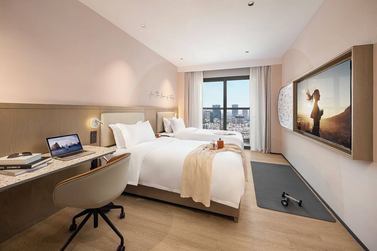 A hotel room with a bed, desk, chair, and a large window with a city view.