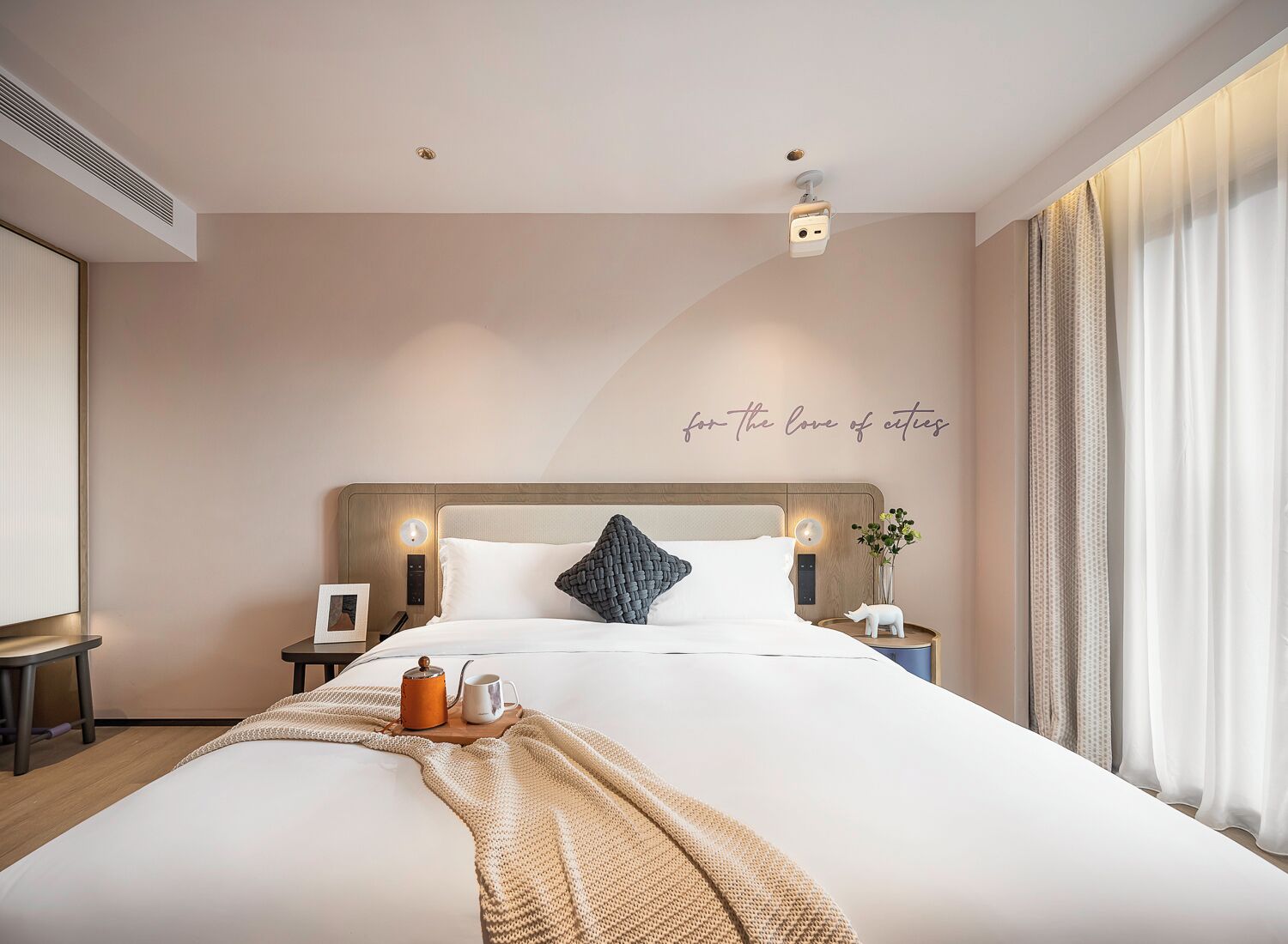 Una habitación de hotel moderna con una cama grande, mesitas de noche, una lámpara de pared y una frase decorativa.