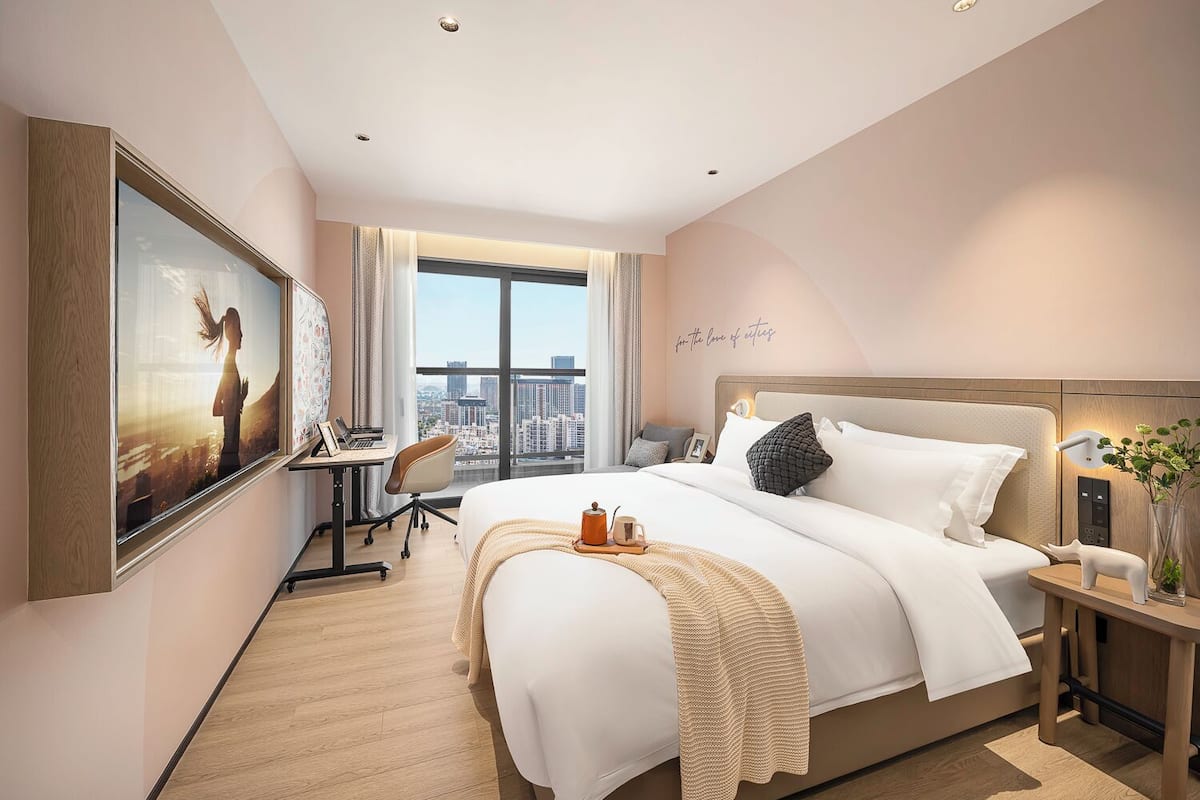 A modern hotel room with a large bed, a desk with a laptop, a view of the city, and a large window.