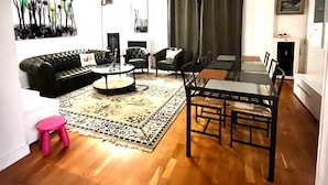 A living room with a black leather sofa, a round coffee table, a dining table with chairs, a patterned rug, and a ceiling fan.