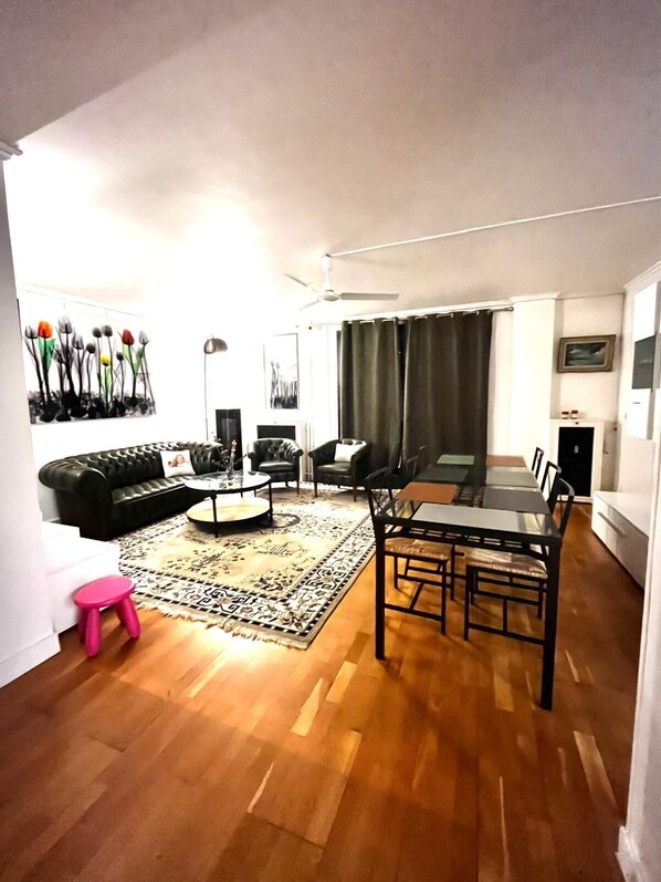 Una sala de estar con un sofá de cuero negro, una mesa redonda de centro, una mesa de comedor con sillas, un tapete estampado y un ventilador de techo.