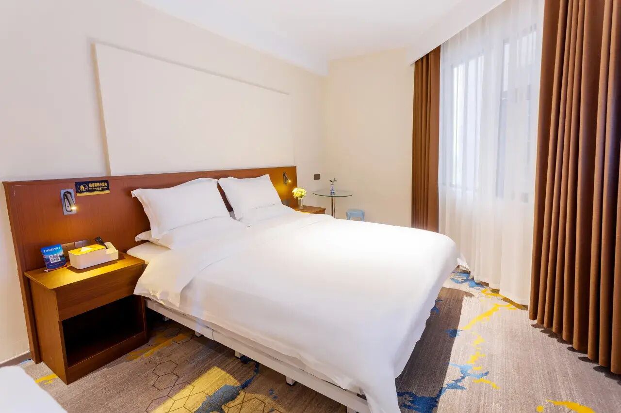 Een hotelkamer met een bed, nachtkastje, lamp, klein tafeltje en een raam met gordijnen.