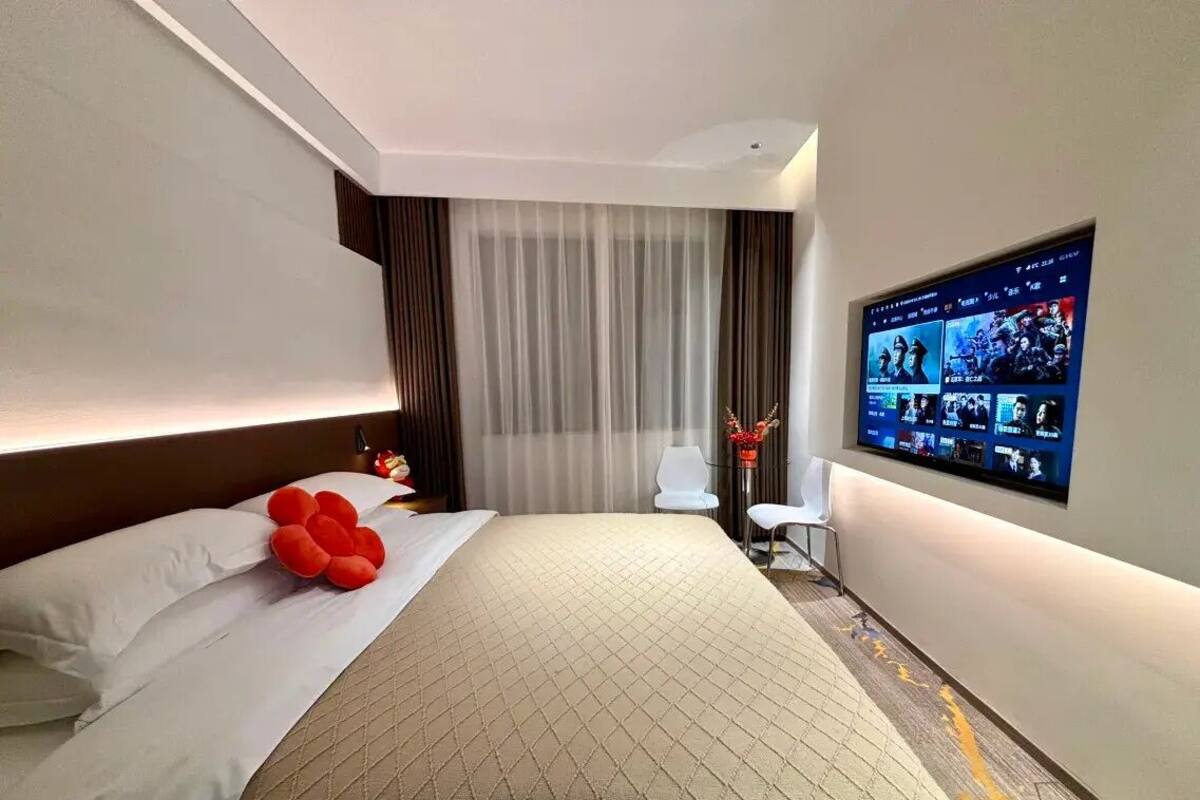 A hotel room with a large bed, a flat-screen TV mounted on the wall, a small table with a chair, and a window with curtains.