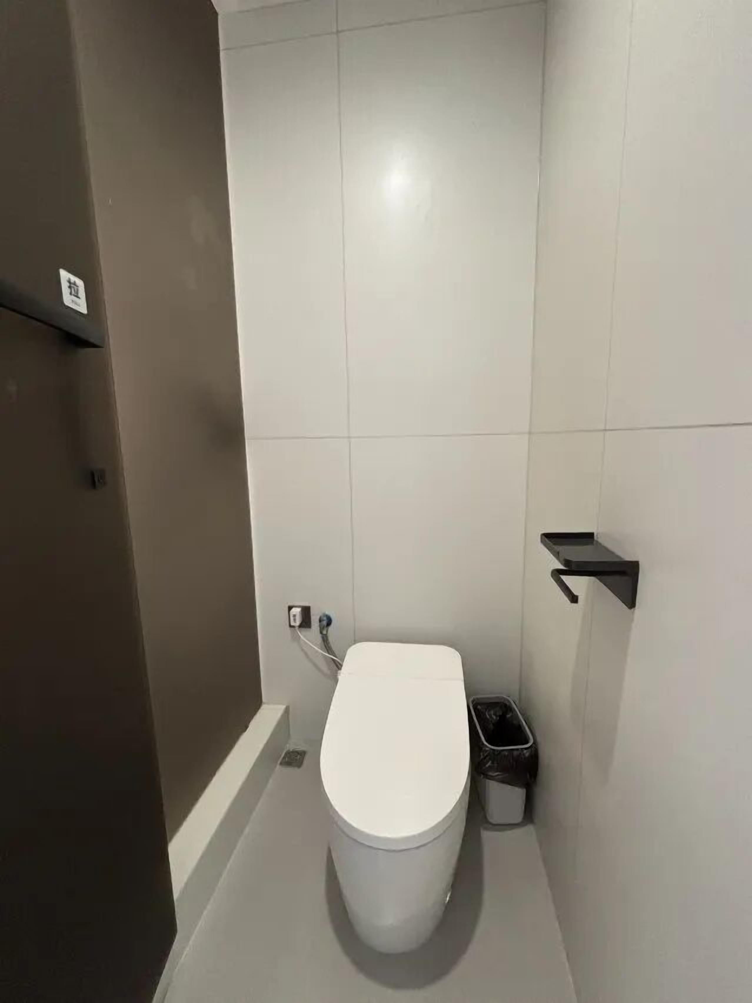 Et lille, moderne badeværelse med et hvidt toilet, en sort skraldespand og en sort hylde på væggen.