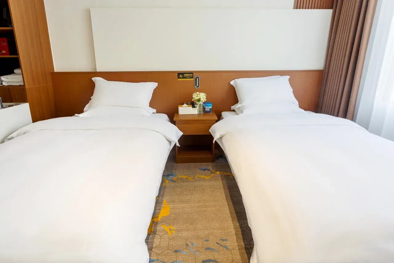 Een hotelkamer met twee eenpersoonsbedden, een houten hoofdbord, een nachtkastje met een bloevas en een bordje met het kamernummer.