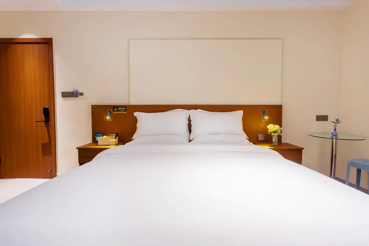 Une chambre d’hôtel avec un grand lit, une tête de lit en bois, des tables de chevet avec des lampes, une petite table ronde et un tabouret bleu.