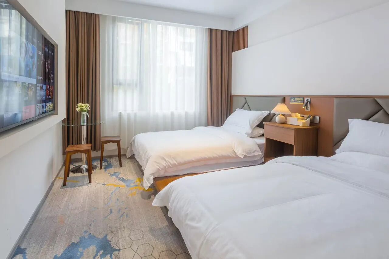 Habitación de hotel con dos camas, televisión, una mesita con un florero y ventana con cortinas.