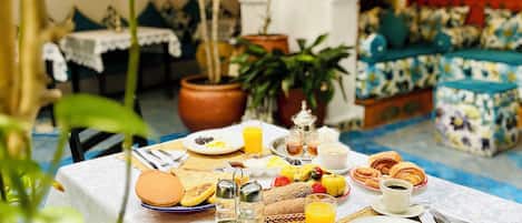 Mesa de desayuno dispuesta con diversos alimentos y bebidas, rodeada de plantas en macetas y un rincón de estar acogedor con cojines.