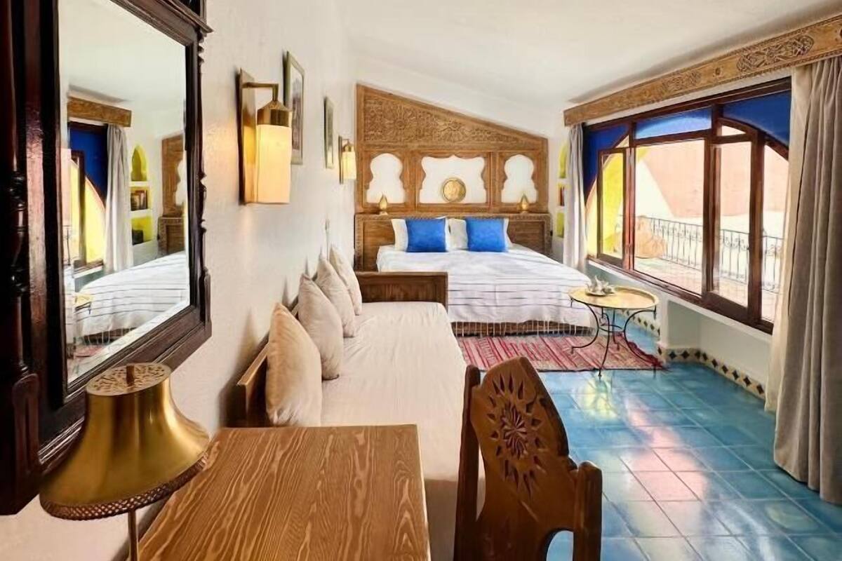 A hotel room with a wooden bed, a wooden table with a lamp, a large mirror, and a view of the outside through a window.
