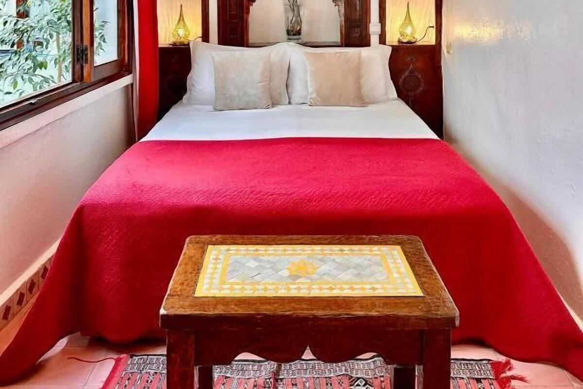 A bedroom with a large bed, red bedding, a chandelier, wooden headboard, and a small table with a patterned tablecloth.