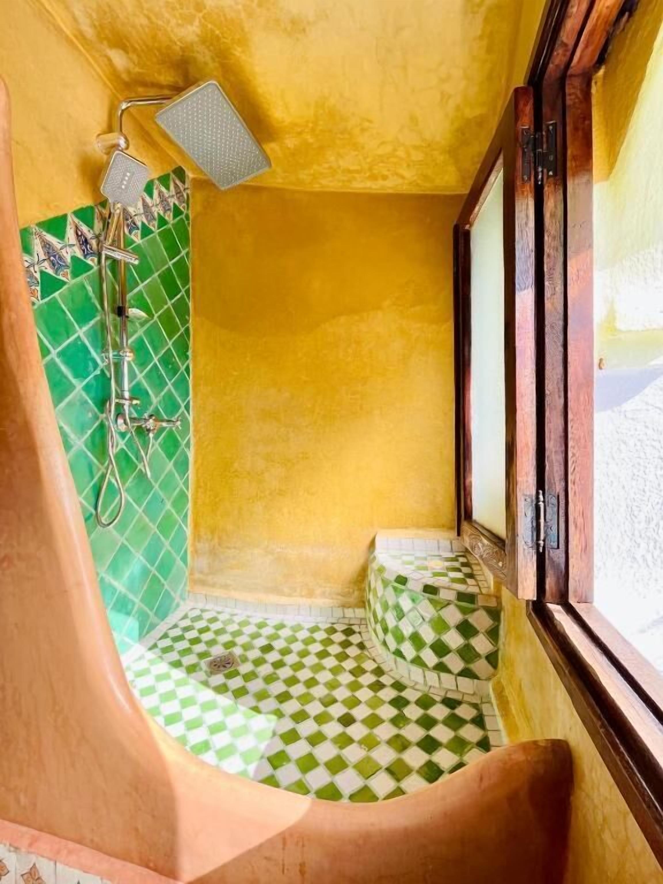 Ein kleines, außergewöhnlich gestaltetes Badezimmer mit einem grün-weißen Schachfußboden, einer Dusche inklusive Handbrause und einem Fenster mit Holzrahmen.
