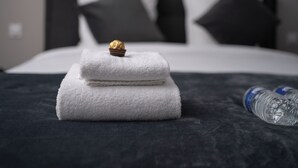 A neatly made bed with folded white towels and a chocolate truffle on top, a water bottle beside it, and a dark-colored headboard with pillows. - Hôtel Sofiane (Paris)