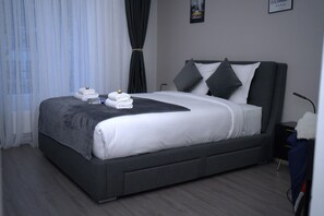 A modern hotel room with a large bed, grey bedding, and a dark grey headboard. There are two pillows on the bed, a small bedside table, and a lamp. - Hôtel Sofiane (Paris)