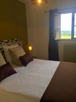 A neatly made bed with a patterned bedspread, a brown headboard, and several pillows. There is a bedside lamp, a window with a view of greenery, and a radiator on the wall. - LARGE BEARNAISE COUNTRY HOUSE (SAUVELADE)