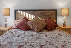 Uma cama com colcha florida, vários travesseiros decorativos e duas luminárias de cabeceira sobre mesas de madeira.