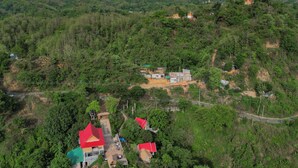 Vista aerea di una zona rurale con diverse case, alcune con tetti rossi, circondata da una vegetazione lussureggiante e colline sullo sfondo.