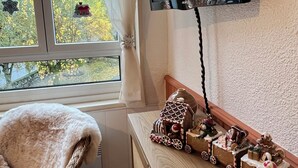 Una habitación con un ventanal grande, un televisor de pantalla plana montado en la pared, una silla de mimbre con una manta blanca y una mesa de madera con una maqueta de una casa de jengibre.