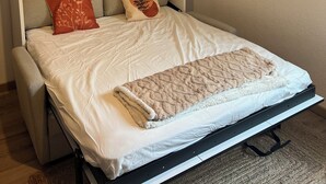 Una cama bien hecha con edredón blanco y dos almohadones decorativos, cabecera con diseños circulares y un cartel en la pared.