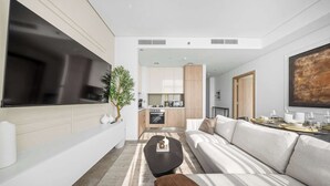 Una sala moderna con un sofá, una mesa de centro y un televisor. Se ve una cocina al fondo con armarios de madera y un comedor con una mesa preparada para dos personas.