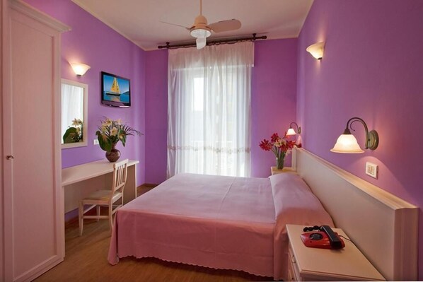 A room with a bed, a desk, a chair, a ceiling fan, a window with curtains, and wall-mounted lamps. - Hotel Plaza Gabicce Mare (Gabicce Mare)