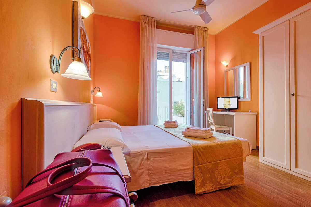 Une chambre d’hôtel avec un lit, un bureau avec un ordinateur, une fenêtre avec des rideaux et un ventilateur de plafond.