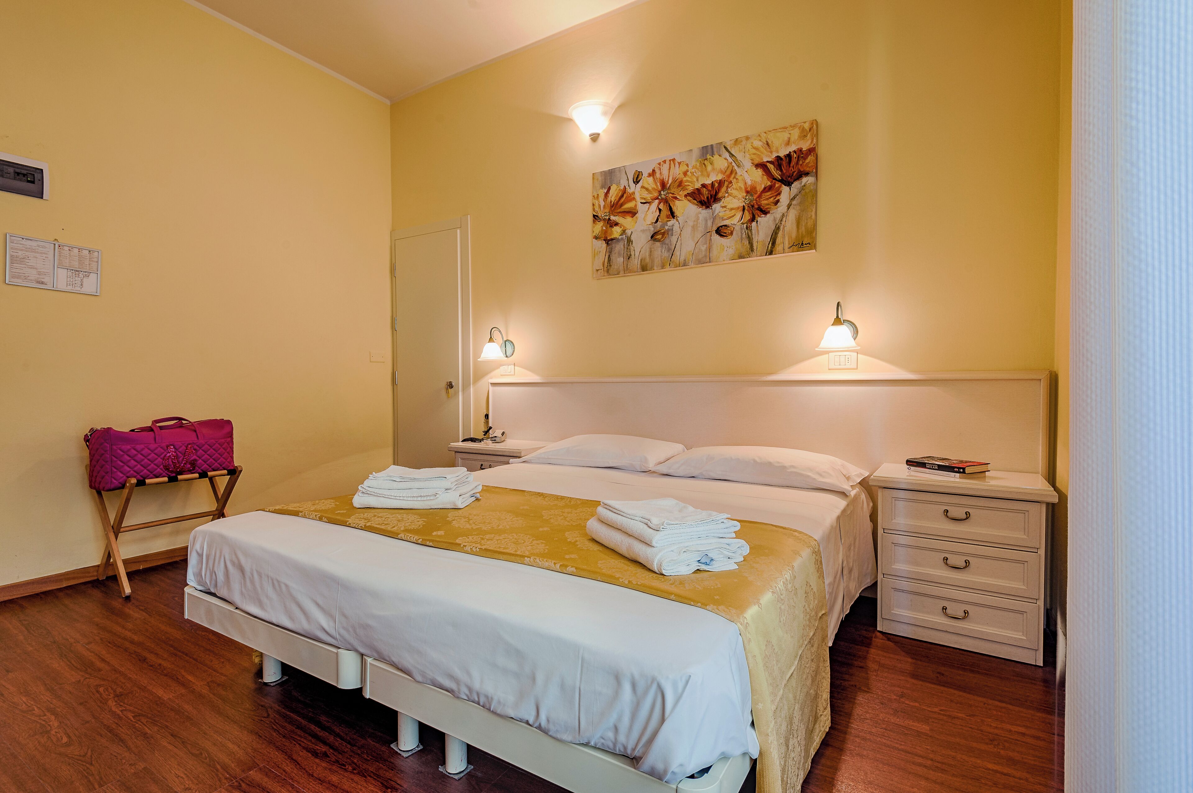 A hotel room with a bed, a bedside table, a nightstand, a lamp, a painting on the wall, and a pink bag on a stand.