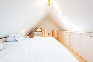 Ein Schlafzimmer mit schrägen Wänden, einem Bett mit weißer Bettwäsche, einem Nachttisch aus Holz, einem Stuhl und einem Kleiderschrank.