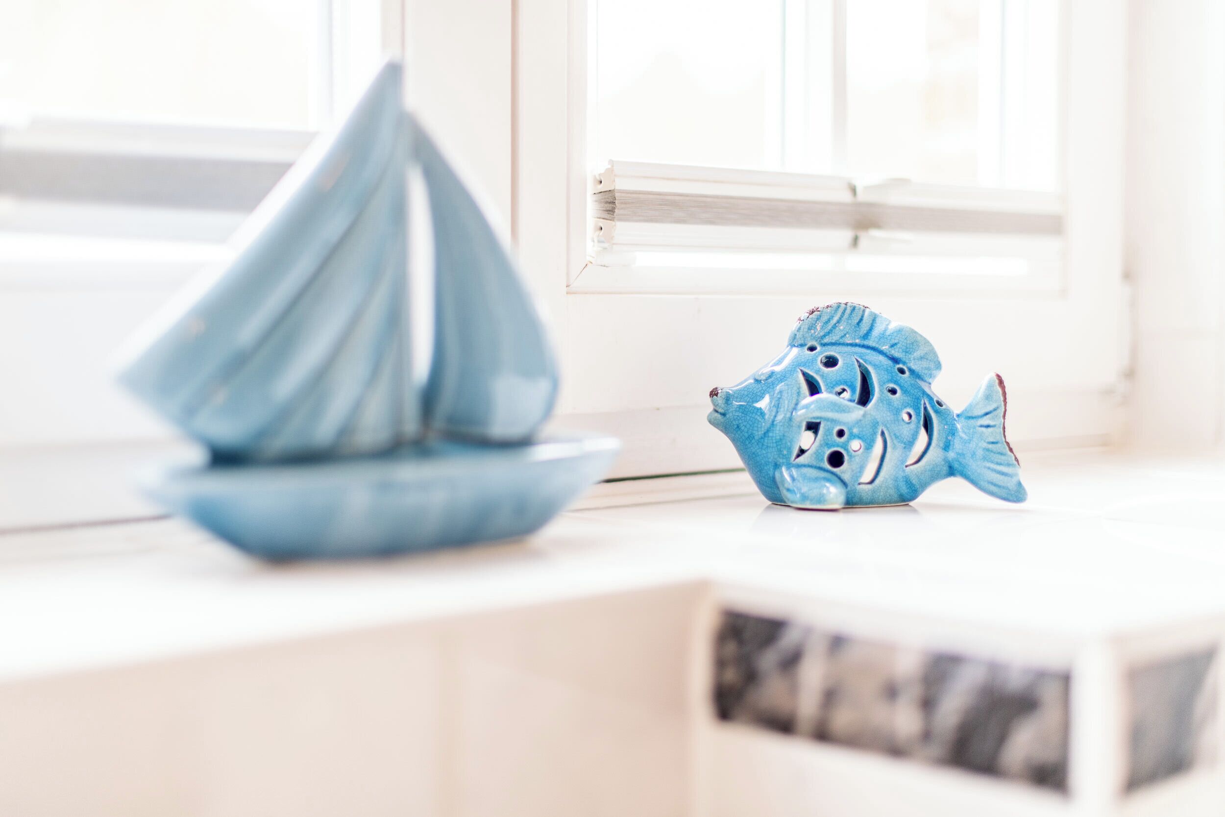 A sailboat and a fish-shaped ornament on a white surface near a window.
