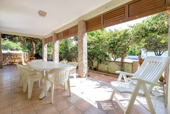 Una terrazza esterna con tavolo e sedie bianche, pilastri in pietra e vista su alberi e una strada.