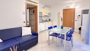 Un espace de vie compact comprenant un coin cuisine, un coin repas avec une table en verre et des chaises bleues, ainsi qu’un canapé bleu avec des coussins blancs.