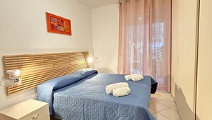 Une chambre à coucher avec une tête de lit en bois, une couvre-lit matelassée bleue, deux serviettes enroulées posées sur le lit, une table de chevet avec une lampe et une fenêtre ornée de rideaux légers.