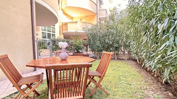 Une table en bois avec quatre chaises, un vase rempli de fleurs et une plante en pot, le tout dans un décor extérieur avec un bâtiment en arrière-plan.