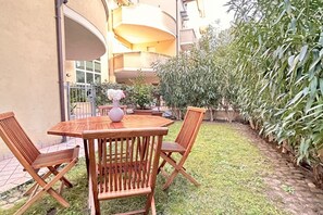 Una mesa de madera con cuatro sillas, un florero con flores y una planta en maceta, en un entorno exterior con un edificio al fondo.