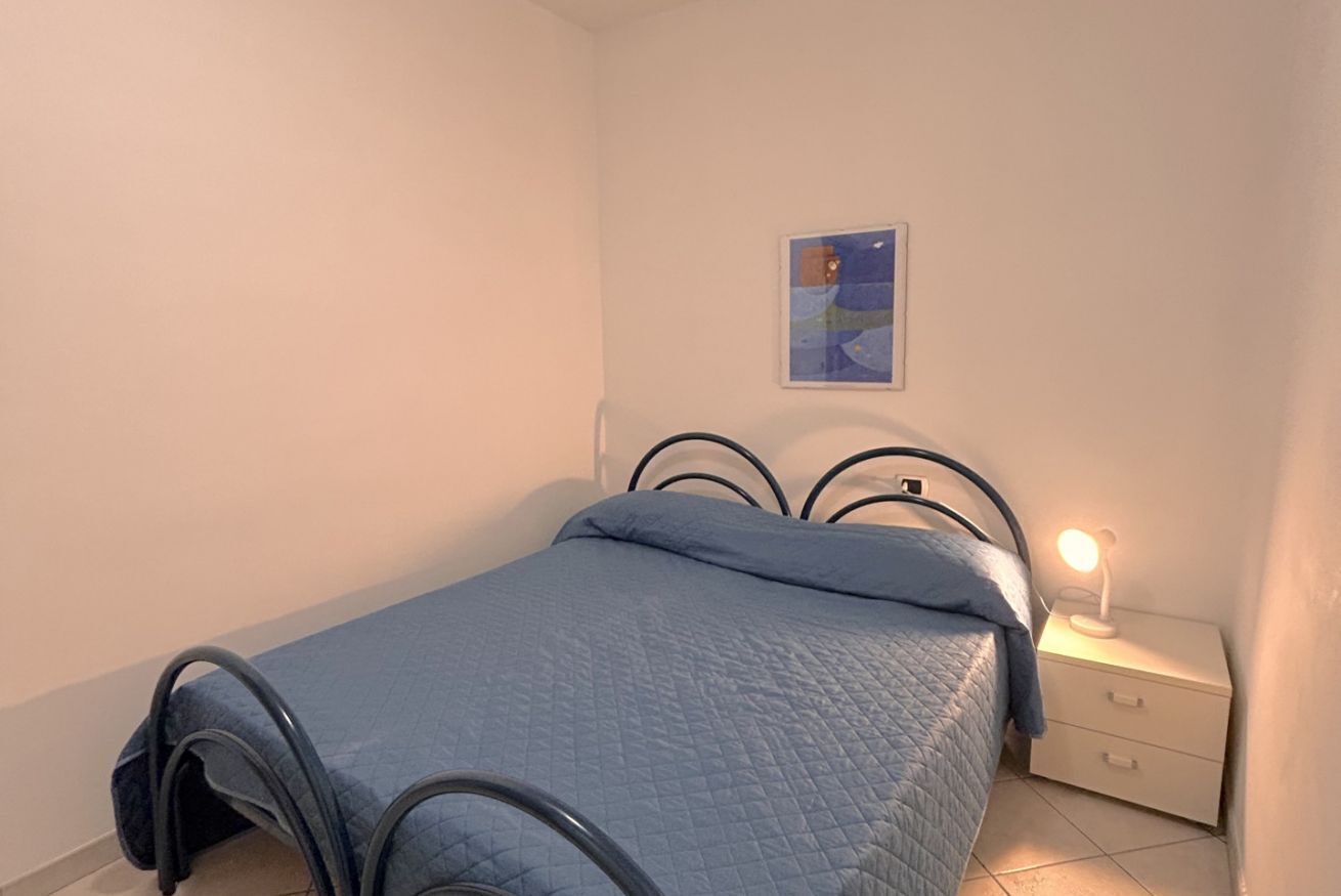 Una cama individual con un edredón azul, una mesita de noche blanca con una lámpara, y un cuadro en la pared.
