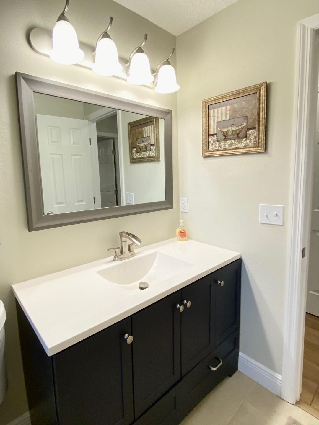 A bathroom with a white sink, dark cabinets, a large mirror, and a framed picture of a bathtub.