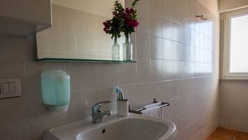 Un bagno con un lavandino bianco, uno scaffale in vetro con vasi e un dispenser per sapone.