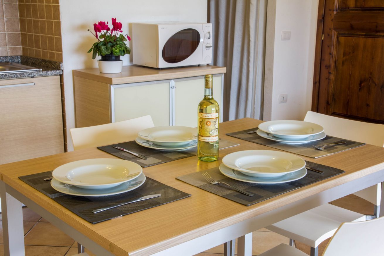 A dining table set with plates, cutlery, and a bottle of wine, with a microwave and potted plant in the background.
