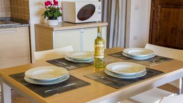 A dining table set with plates, cutlery, and a bottle of wine, with a microwave and potted plant in the background.