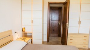 A bedroom with a bed, bedside table, lamp, wardrobe, and a door leading to another room. - GF Apartments La Vigna - Vigna Garden - SCIROCCO (Badesi)