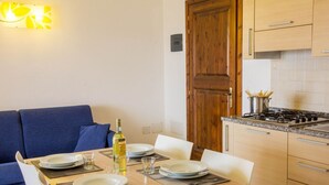 A dining area with a wooden table set for four, a blue sofa, and a kitchen with a stove and cabinets. - GF Apartments La Vigna - Vigna Garden - SCIROCCO (Badesi)