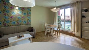 Un salon comprenant un canapé, une table à manger avec des chaises, une table basse, une grande fenêtre avec des rideaux et un luminaire au plafond.