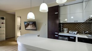 Une cuisine moderne avec des comptoirs blancs, des armoires en bois foncé, un four encastré et un grand îlot central doté d’un évier. Des luminaires suspendus sont placés au-dessus de l’îlot, et un téléviseur à écran plat est fixé au mur.