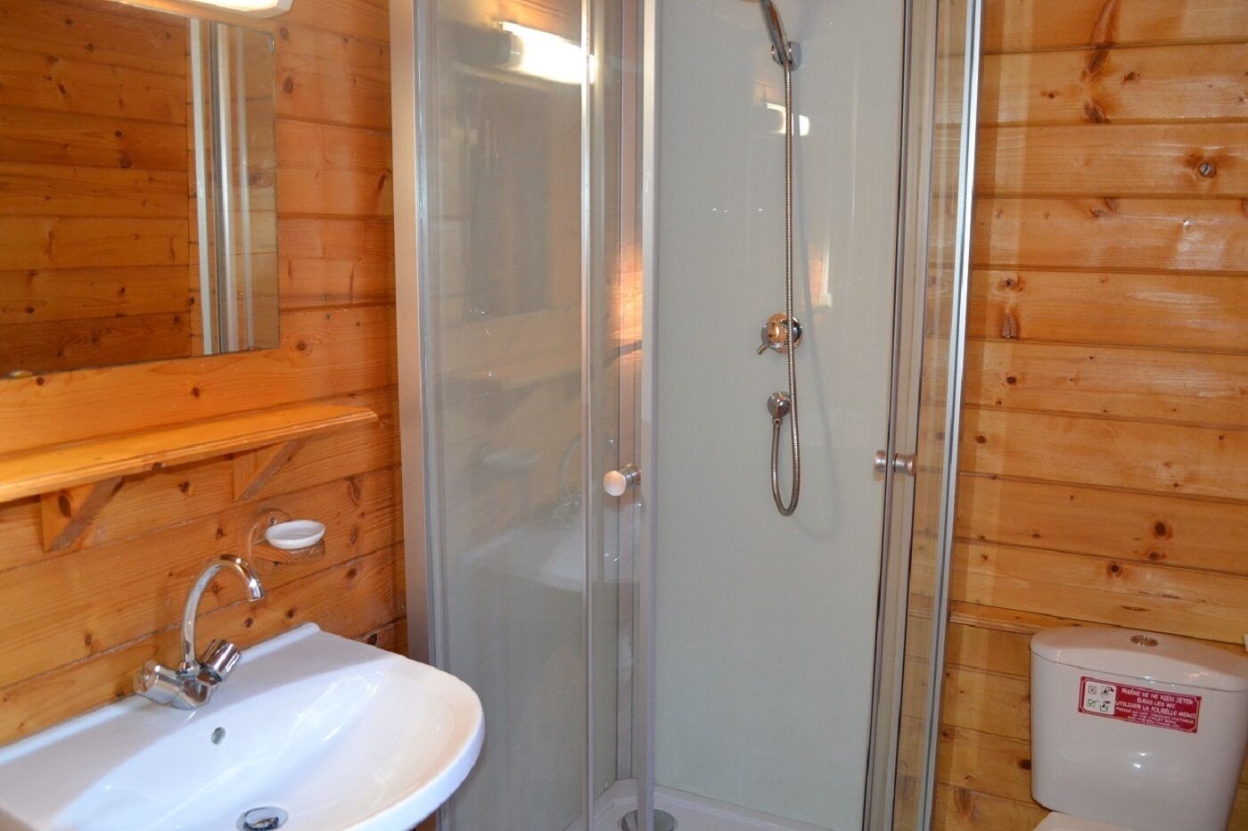 Et badeværelse med bruseniche i glas, håndvask med kromarmatur og toilet med rød klistermærke.