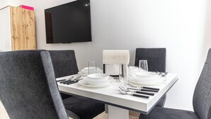 Un comedor moderno con una mesa blanca para cuatro, sillas grises tapizadas, un televisor de pantalla plana en la pared y un mueble de madera con detalles rosados.