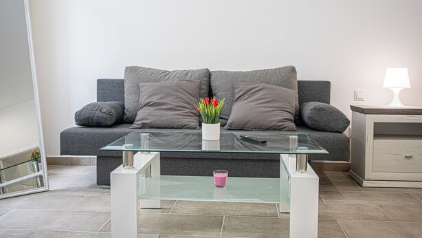 A modern living room with a grey sofa, a glass coffee table, and a bedside table with a lamp. - Beautiful 1BR Apartment For Long Stays (Vienna)