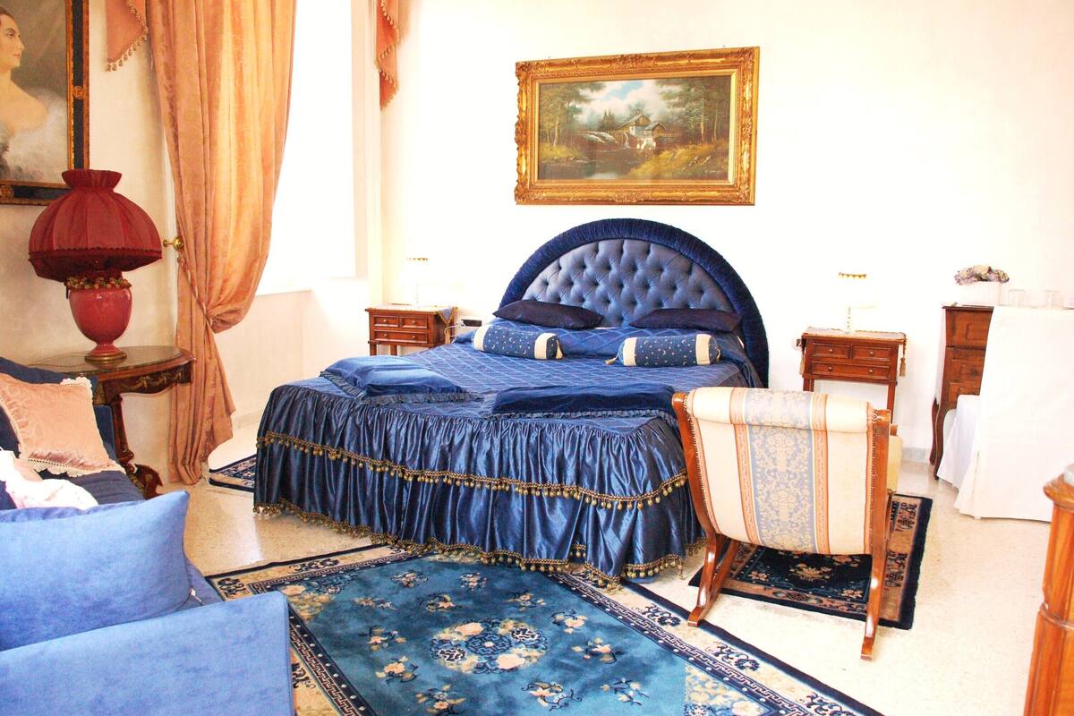 Un dormitorio con una cama grande, una silla, una mesita de noche, un aparador y un cuadro en la pared.