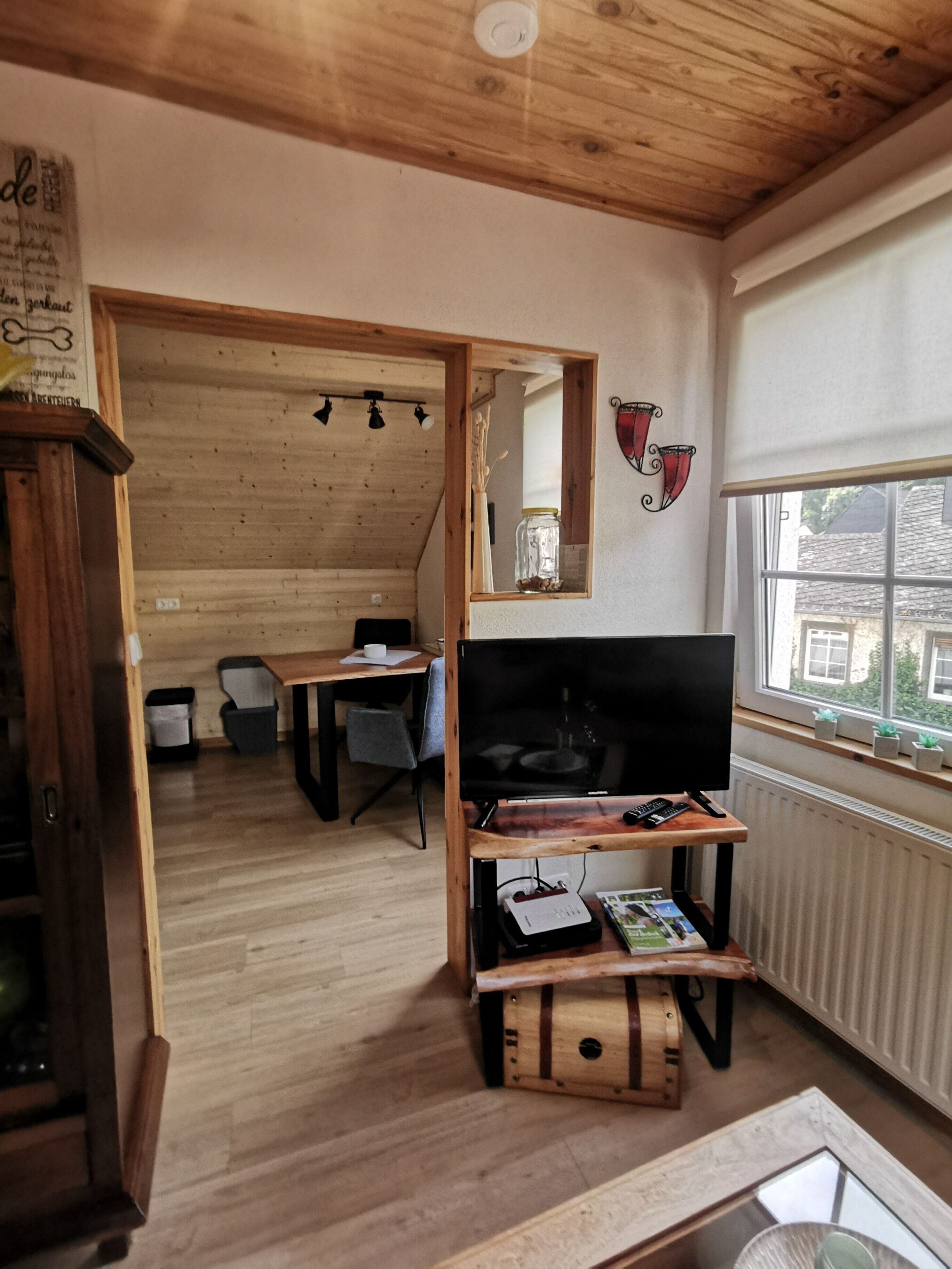 A cozy room with a wooden ceiling, a TV on a wooden stand, a window with a view, and a mirror on the wall.