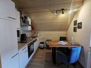 A compact kitchen with a dining area, wooden ceiling, and a small trash bin.