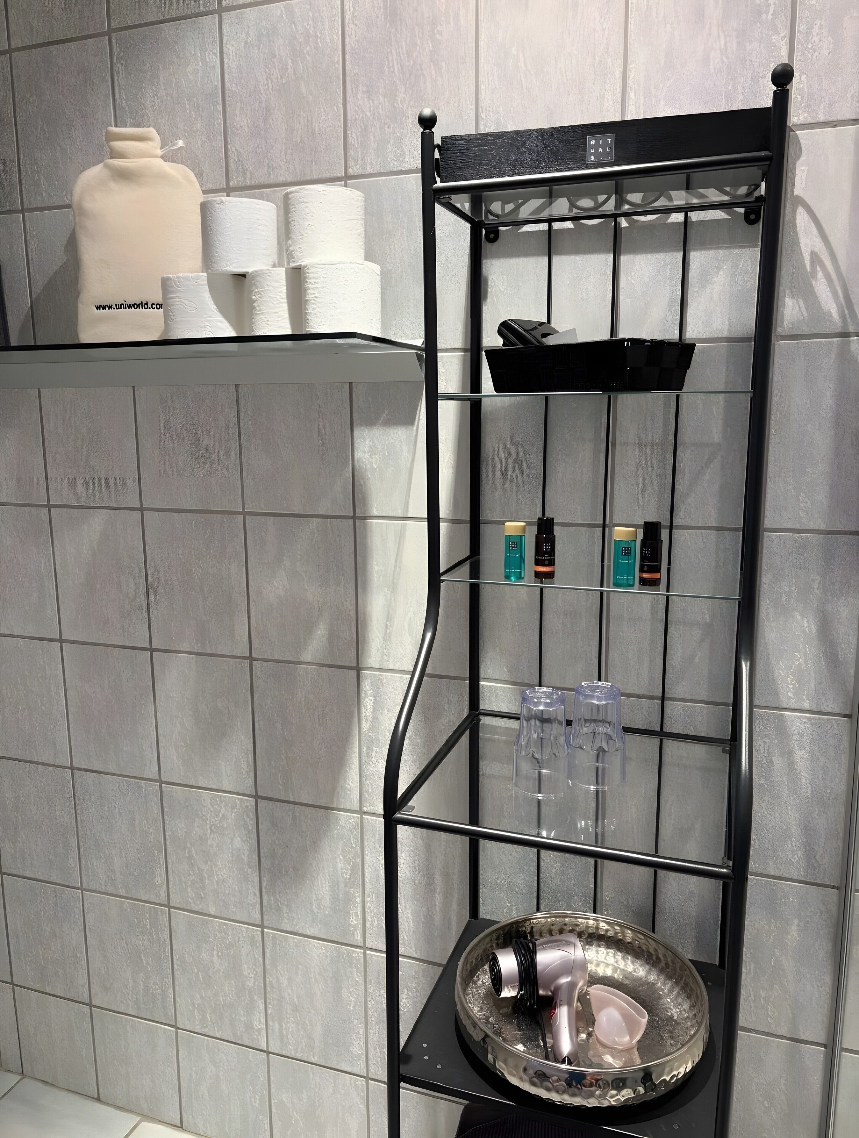 Ein Badezimmer mit einem schwarzen Metallregal, das Toilettenartikel beherbergt, einer Glasbowl mit einem Fön und einer gefliesten Wand.