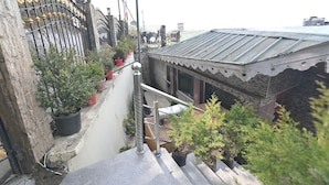 Un stretto vicolo con una scala metallica che conduce a un edificio con tetto in lamiera ondulata. Lungo le pareti si trovano piante in vaso e un recinto metallico con elementi decorativi.